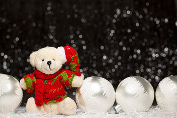 Christmas view of teddy bear in red green sweater and Christmas hat and four silver evening balls with white snow on it on dark background with silver bokeh. Holiday concept with copy space