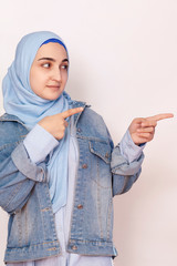 Cute Muslim woman with cheerful face expression points away with her forefinger, showing to copy space on the corner. Portrait of Muslim prayer woman in hijab