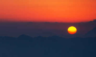 The disk of the Sun sets on the silhouette of mountains with a warmth horizon