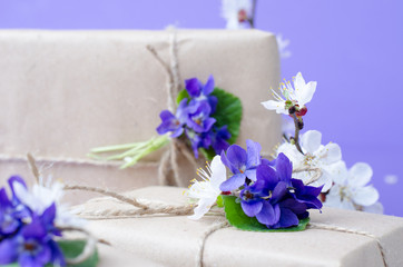 Beautiful gift boxes wrapped in simple brown craft paper decorated with live flowers of violet on a violet background.