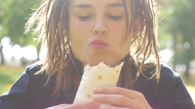 Young hipster girl with dreadlocks eats kebab. Girl eats fast food shawarma in the park. Sunny day.