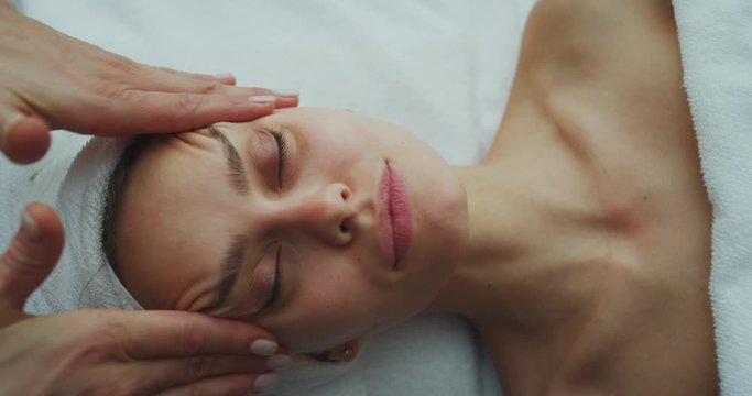 Close up of a beautiful young smiling woman with a towel on her head is receiving a facial massage and spa treatment for perfect skin in a luxury wellness center.