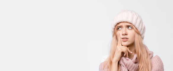 Obraz na płótnie Canvas Studio portrait of attractive blonde girl in white hat and pink sweater. Worry female presses a finger to her cheek and thinks about problems in life. Emotions concept. Copy space.