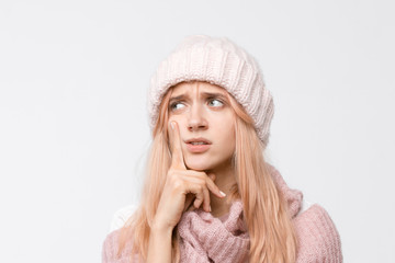 Studio portrait of attractive blonde girl in white hat and pink sweater. Worry female presses a finger to her cheek and thinks about problems in life. Emotions concept.