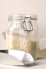 White uncooked, raw long grain rice in glass storage jar with metal scoop on white kitchen table