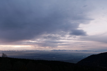 Winter landscape at sunrise. Silhouette of hills and trees in the foreground and snow capped mountain range of the European alps in the background.