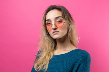 Romantic blonde-haired girl in trendy heart glasses posing with shy smile. Studio close-up portrait on a pink background.