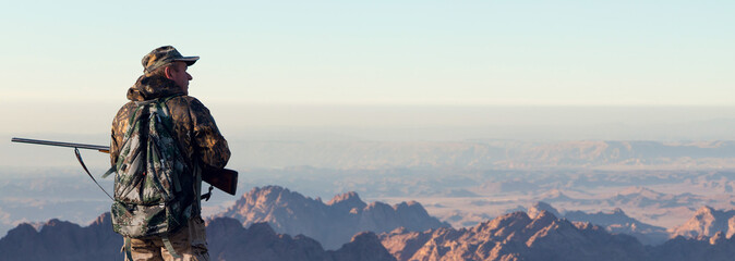 A hunter in camouflage and a gun in his hands in the early morning on top of a mountain stands...