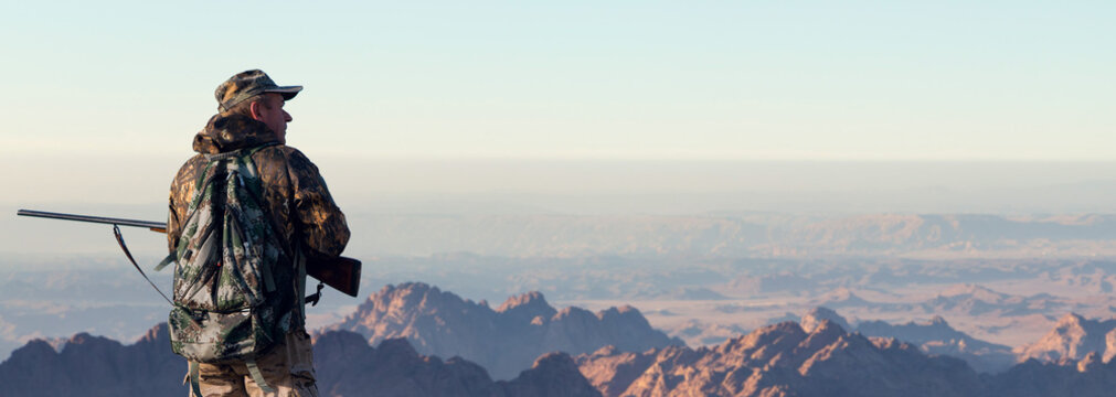 A hunter in camouflage and a gun in his hands in the early morning on top of a mountain stands looking for a target. Beautiful mountain landscape, the opening of the hunt.