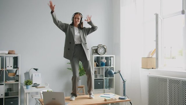 Beautiful office worker in suit is dancing on desk in office wearing headphones enjoying break from work. Crazy people, workplace fun and lifestyle concept.
