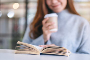 Closeup image of a woman holding and reading a book while drinking coffee on wooden table