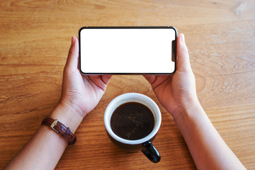Top view mockup image of hands holding black mobile phone with blank screen with coffee cup on wooden table