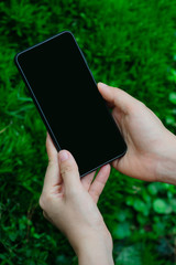 Close up woman hand holding and touching screen on the modern black smartphone mock up in vertical position with blank screen against blurred grass background. Clipping path inside.