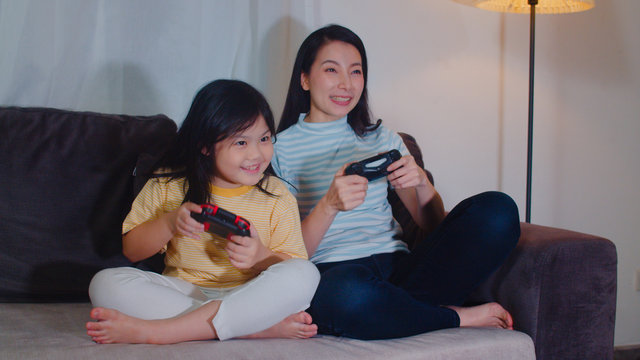 Young Asian Family And Daughter Play Games At Home In Night. Korean Mother With Little Girl Using Joystick Funny Happy Moment Together On Sofa In Living Room. Funny Mom And Lovely Child Are Having Fun