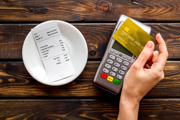 Electronic payments in restaurant. Hand bring bank card to terminal near bill on dark wooden...