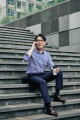 Handsome young Asian man in glasses sitting on steps, drinking take away coffee and calling on phone