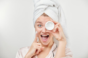Excited girl with towel on head holding jar of facial hydrating cream by eye