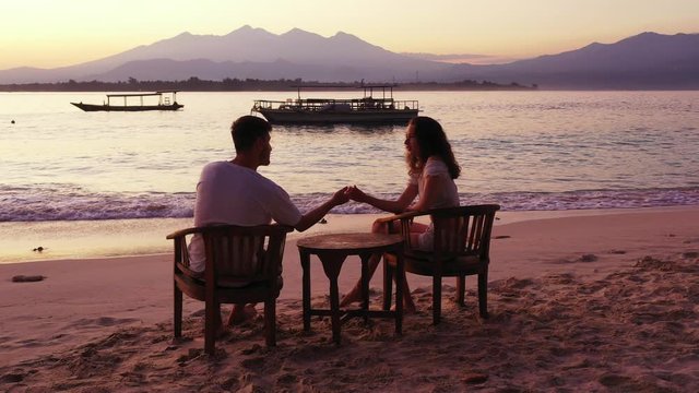 Bora Bora Island - Couple having a romantic moment while enjoying the sunset by the shoreline of the sandy beach with mountain stretch and boats floating on the background - Panoramic Shot 