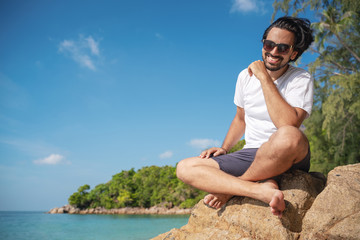 Naklejka na ściany i meble Young fashionable stylish handsome latin man in a white t-shirt and sunglasses on a bright tropical beach, vacation and travel to tropical countries