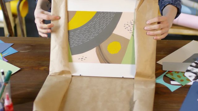 Close up hands of female decorator standing by desk and wrapping framed picture or collage in paper
