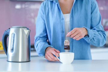 Woman putting tea bag in a cup and using an electric kettle for brewing hot tea at home at kitchen