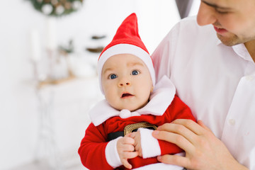 a baby in a little Santa suit in dad's arms. The father looks at him and enjoys his fatherhood and is ready to celebrate Christmas and New Year