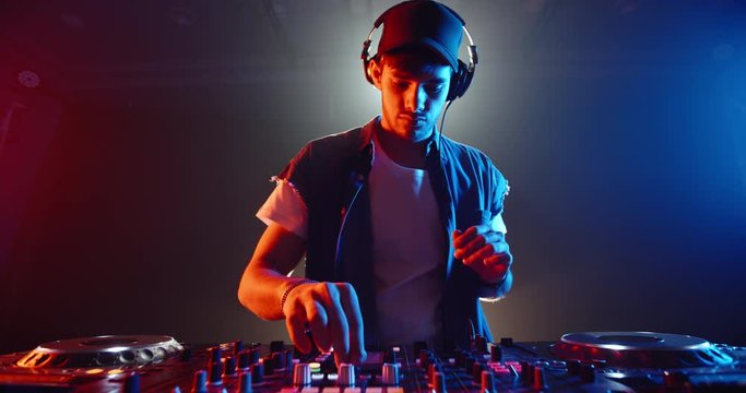 Cool dj standing at a mixer controller composing a dance music list, dancing while spotted by nightclub colorful lights - nightlife concept 4k footage