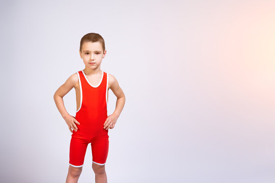 Portrait of a little cheerful boy in a red wrestling tights holds his hands on his sides, looks confidently at the camera and poses on a white isolated background. concept of a little fighter athlete