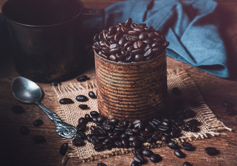 Dark roasted coffee beans in a rusty can on burlap and wood table. Dark mood coffee beans photography.