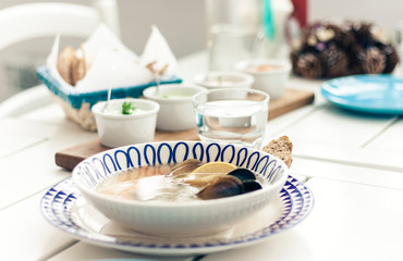 Seafood soup with mussels, prawns and fish in white bowl with blue ornament with sauce in small bowls for traditional meze dinner on the wooden table on background.