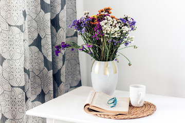 bouquet of flowers, open book and glasses, lemon tea in a white ceramic cup on a wicker stand on the table, cozy home interior.