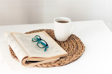 open book and glasses, lemon tea in a white ceramic cup on a wicker stand on the table, cozy home interior.