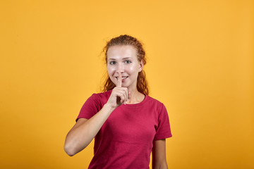 Pretty young lady in red shirt keeping hand on on mouth, shh gesture isolated on orange background in studio. People sincere emotions, health concept.