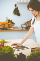 Young woman looking for a new recipe for cooking in a kitchen. Housewife is making online shopping by tablet computer and credit card