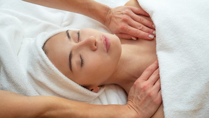 Close up portrait of a beautiful young smiling woman with a towel on her head is receiving a facial massage and spa treatment for perfect skin in a luxury wellness center.