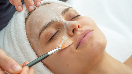 Close up of cosmetologist applies facial clay mask on an young beautiful woman face before ultrasonic cleaning procedure for skin pores and deep moisturizing.
