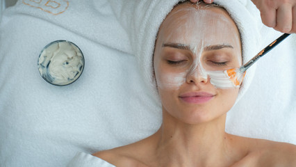 Close up of cosmetologist applies facial clay mask on an young beautiful woman face before ultrasonic cleaning procedure for skin pores and deep moisturizing.