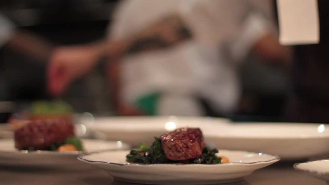 Dolly, closeup shot of steaks being plated while a chef sprinkles salt in the background in a kitchen of a high-end restaurant.