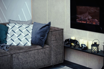 The gray velor sofa in the dark loft room has a bright light from the eternal light and an artificial fireplace. Inner attic with concrete walls and a decorated Christmas tree with gift boxes.