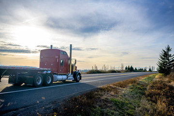 Big classic rig red semi truck with flat bed semi trailer running on the evening road with sunset