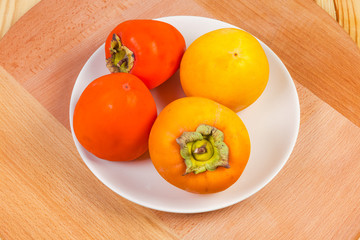 Different persimmon fruits on white dish on the wooden surface