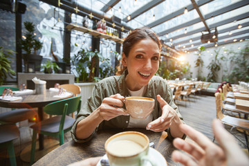Fisheye view of smiling young woman holding coffee cup talking to friend across table in cafe, POV