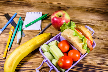 Ripe apple, banana, different stationeries and lunch box with hamburger, cucumbers and tomatoes on wooden table