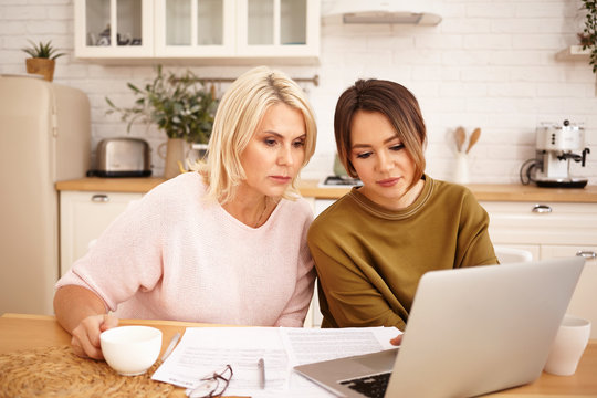 Serious Dark Haired Girl Sitting At Kitchen Table With Her Middle Aged Mother Using Wifi On Generic Portrable Computer, Drinking Coffee And Calculating Family Budget, Planning Expensive Purchase