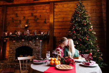 Loving couple sitting in a large wooden house with a fireplace and a Christmas tree, kissing at the holiday table