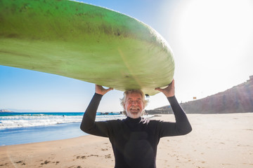 happy senior with surftable on his head is smiling and laughing - old and mature man having fun surfing with a black wetsuits - active retired adult doing activity alone - Powered by Adobe