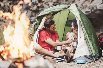Happy trekker couple camping tent with dog next bonfire  - Hipster man and woman having fun mountaineering together - Love relationship and travel lifestyle concept - obrazy, fototapety, plakaty
