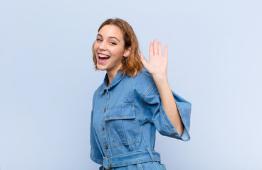 young blonde woman smiling happily and cheerfully, waving hand, welcoming and greeting you, or saying goodbye against flat color wall