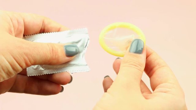 Woman's hand with gray manicure opening condom on pink background.