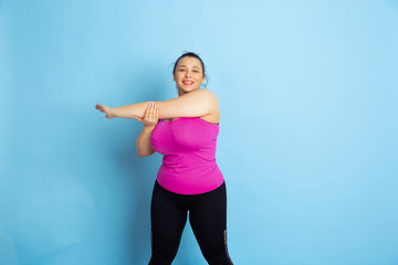 Fototapeta na wymiar Young caucasian plus size female model's training on blue background. Concept of sport, human emotions, expression, healthy lifestyle, body positive, equality. Making stretching exercises.